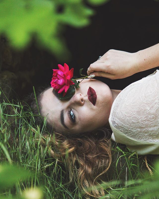 A woman lies in the grass with a red rose on her face in a dreamy, intimate portrait.