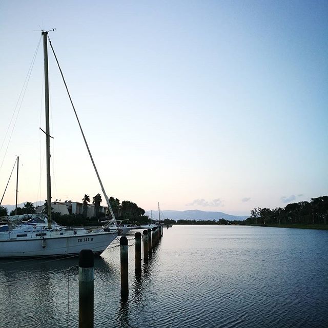 A sailboat is docked in a tranquil harbor at sunset, reflecting the cool blue sky.
