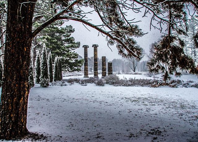 A winter scene with snow-covered ground, trees, and weathered columns standing in a tranquil garden.