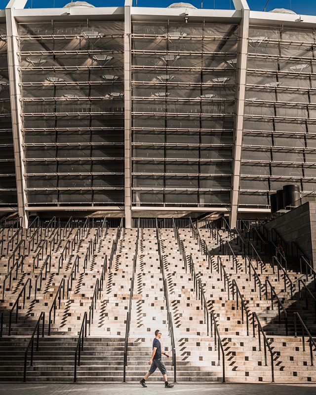 A man walks past a large stadium with many stairs and railings on a sunny day.