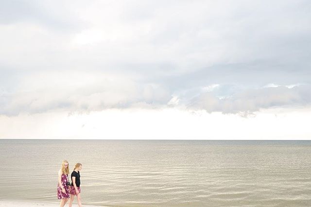 Two women walk along a tranquil beach on an overcast day, enjoying the calm ocean view.