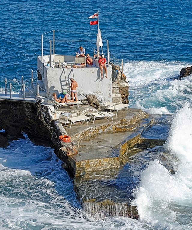 People relaxing by the sea with white waves hitting a rock platform on a sunny day.