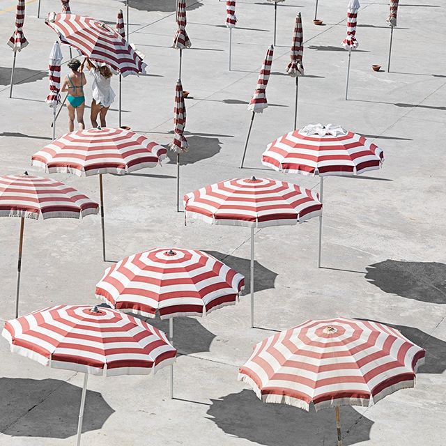 Red and white striped umbrellas cast shadows on a sunny beach scene with two women in the background.