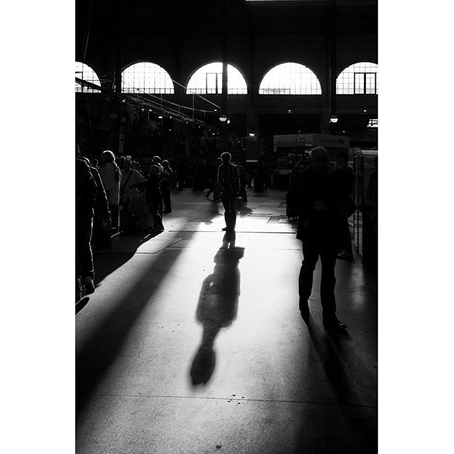 A black and white image capturing people walking in a market hall with strong shadows.