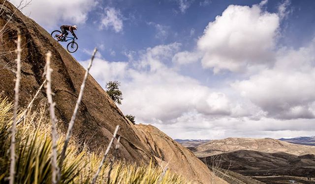 A mountain biker rides down a steep rock face in a rugged outdoor landscape under a partly cloudy sky.