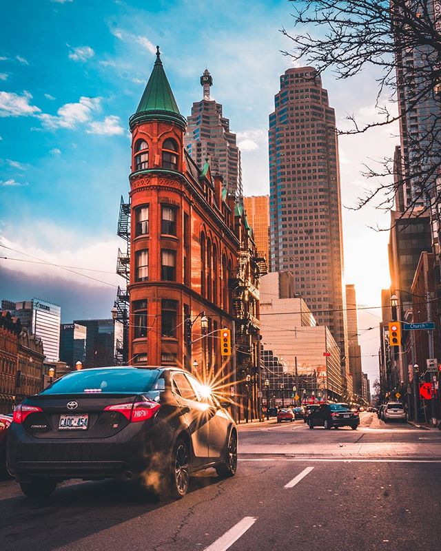 A black car drives on a city street with a historic building and modern skyscrapers under a bright sky.