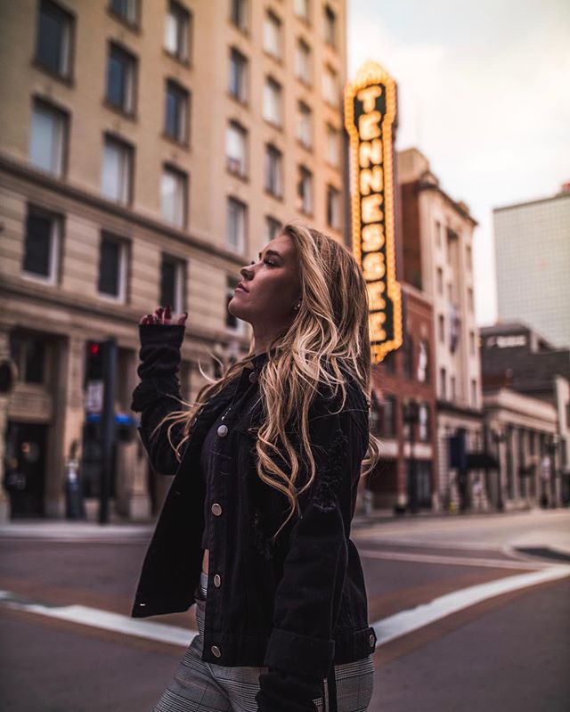 A blonde woman in a jacket stands on a city street looking up at a vintage theater sign.