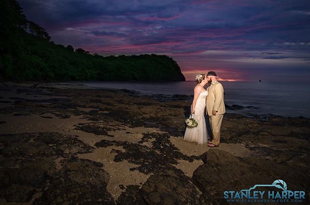 A bride and groom share a kiss on a rocky beach at sunset, perfect for destination wedding and romantic getaway concepts.