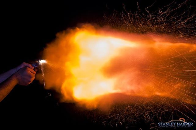 A close-up of a hand holding a gun that is shooting a burst of flame in the dark.