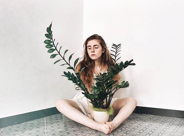 A young woman sits cross-legged with a ZZ plant in a minimalist corner setting, creating a calm, editorial vibe.