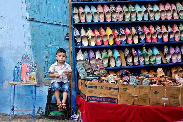 A young boy sits near a shoe stall with colourful slippers and traditional footwear in a Moroccan market.