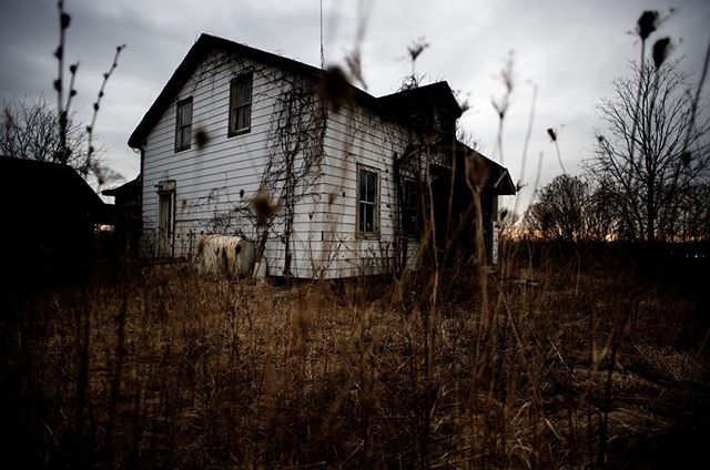 An old, abandoned house stands in a field under a cloudy sky, evoking a sense of mystery and isolation.