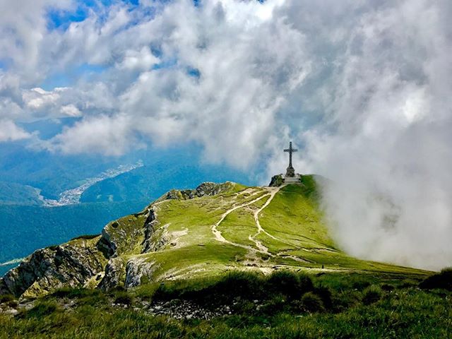 A scenic view of a cross on a mountain peak surrounded by clouds and a winding hiking trail. 