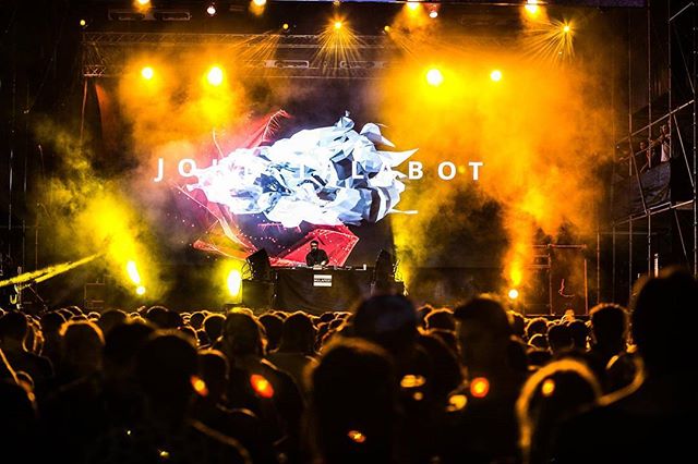 A DJ performs on stage at a concert with a large crowd, bathed in bright yellow lighting and dynamic visuals.