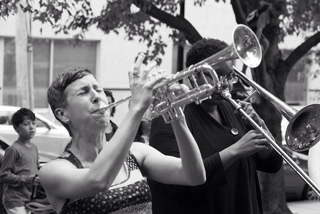 Two musicians play trumpet and trombone at an outdoor event in this black and white candid photograph.
