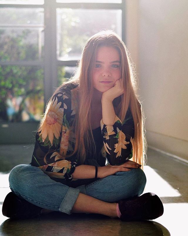 A young woman with long hair sits cross-legged in a sunlit room, looking thoughtful.