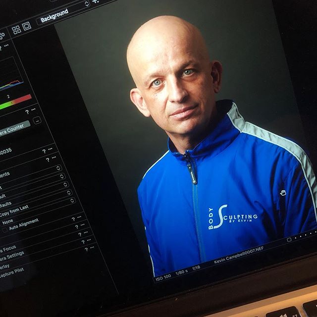 A bald man in a blue jacket poses for a portrait in a studio setting.