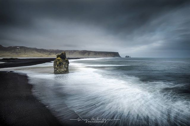 An Icelandic black sand beach with a dramatic rock formation and a blurred ocean under a cloudy sky.