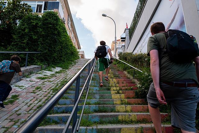 People walk up a colorful, steep staircase in an urban setting. The stairs are painted with rainbow colors.
