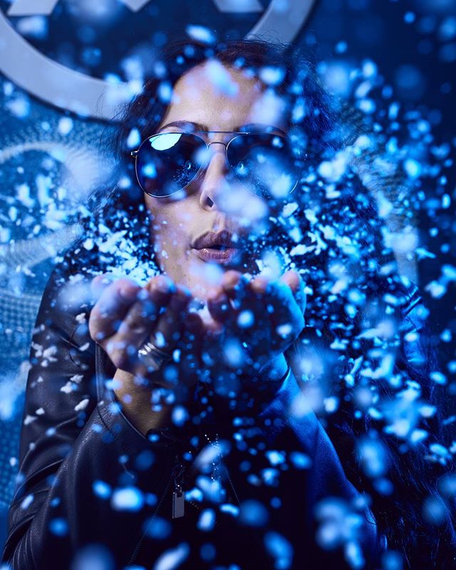 A woman in sunglasses blowing blue powder in a studio environment, creating a mysterious and moody atmosphere.