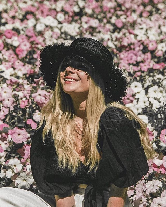 A smiling woman wearing a hat poses among a field of pink and white flowers in this outdoor fashion shot.