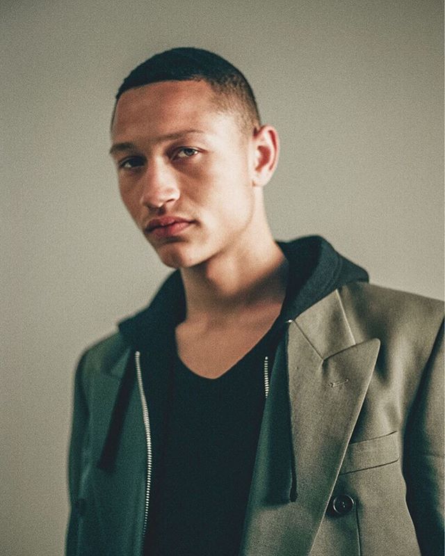 A confident man poses in a blazer and hoodie, showcasing modern urban style in a studio setting.