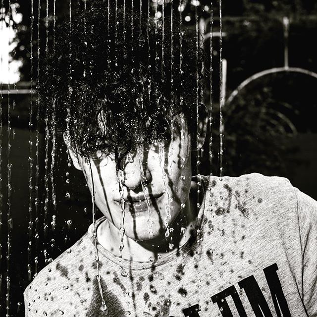 A black and white image of a young man with water streaming down his face and onto his t-shirt.