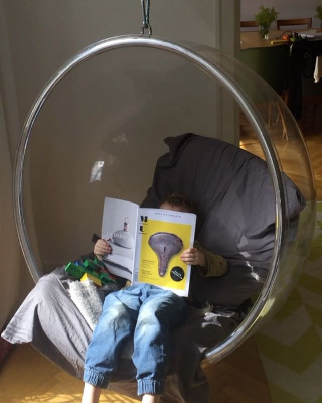 A child sits in a clear bubble chair reading a magazine in a sunlit room at home.