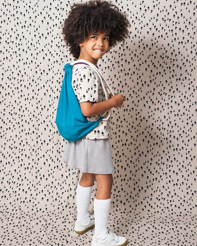 A young girl with curly hair smiles, wearing a backpack and casual clothes in a playful indoor setting.