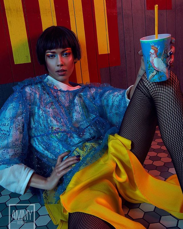 A stylish woman poses with a cockatoo cup, illuminated by vibrant lighting, in a modern editorial fashion shot.