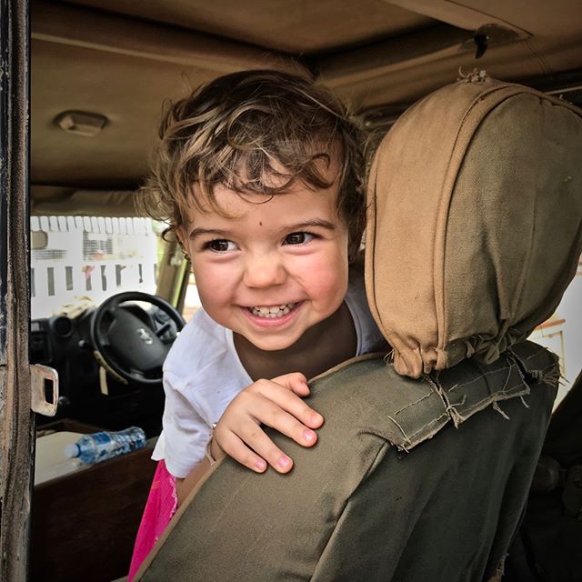 A cute girl smiles from inside a car, looking out the window with a happy expression. 