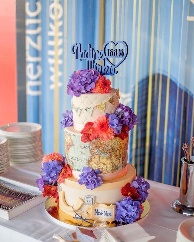 A three-tier travel themed wedding cake decorated with flowers, displayed at a wedding reception.