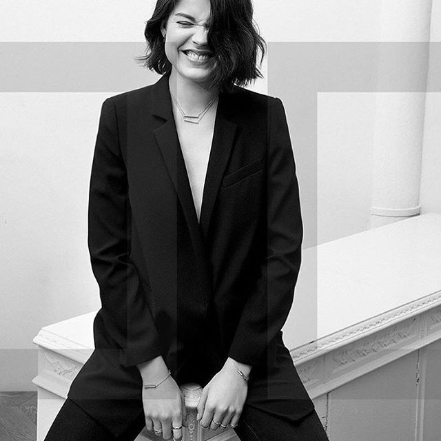 A stylish woman smiles, wearing a black blazer and jewelry in a modern studio setting.