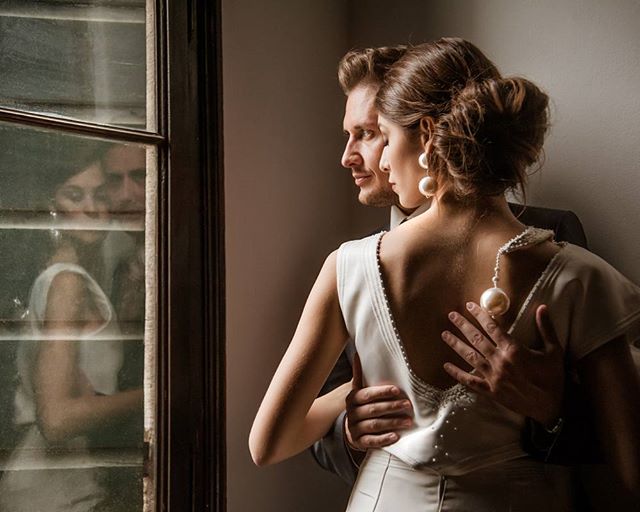 A couple embraces near a window, their reflections visible, evoking a sense of intimacy and romance.