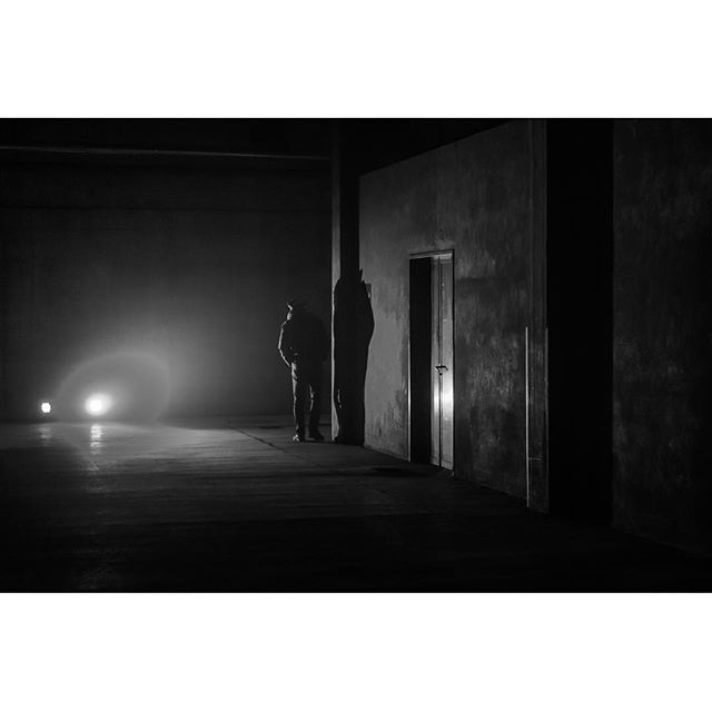 A monochrome image shows two people standing in a dark room with a mysterious and suspenseful atmosphere.
