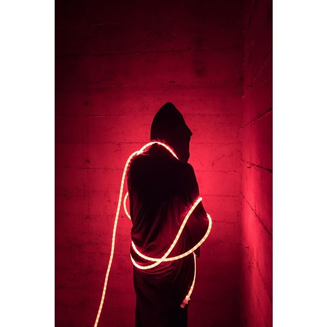 A hooded figure stands in a red room, wrapped in neon light for a mysterious visual.