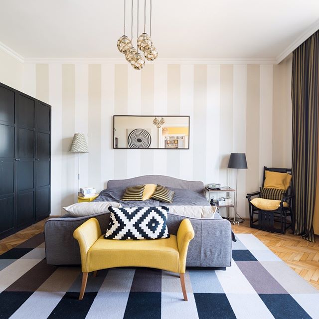 A cozy bedroom features a gray bed, yellow accents, and a striped wall for a modern and relaxing space.
