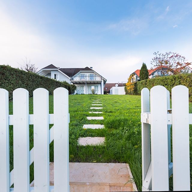 A white picket fence opens to a lush green lawn leading to a modern house under a bright blue sky.