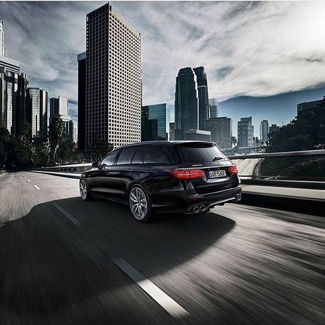 A black Mercedes-AMG estate car speeds down a highway with a cityscape backdrop for luxury car advertising.