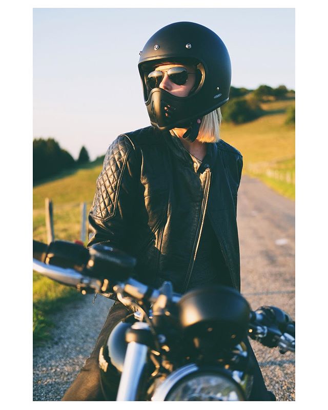 A woman in a helmet and leather jacket poses with her motorcycle on a rural road.