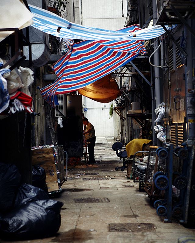 A man stands in a narrow urban alleyway with awnings, garbage, and a gritty city atmosphere.