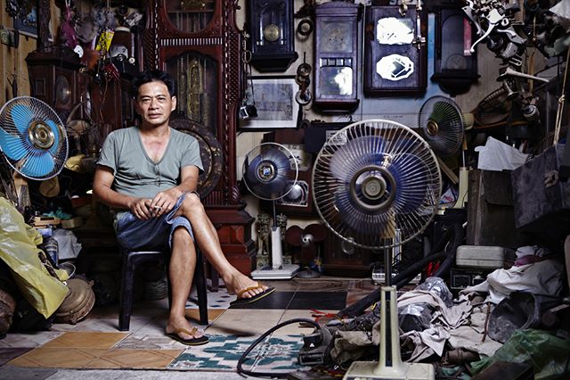 Man sits among clocks and fans in a cluttered workshop, capturing a moment of calm amidst the chaos.