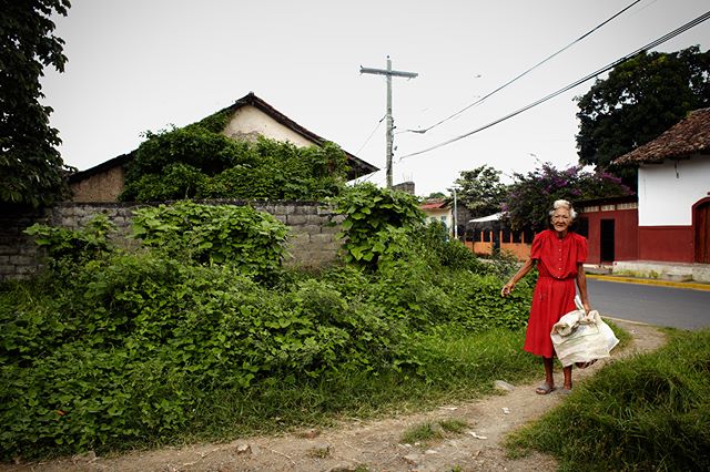 An elderly woman in a red dress walks along a village street on a cloudy day.