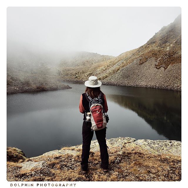 A woman with a backpack and hat stands on a mountain overlooking a calm lake in a misty landscape.