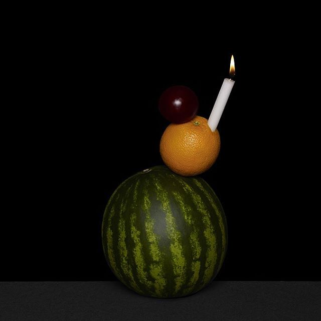 A still life shot features a watermelon topped with an orange, plum, and lit candle on a dark background.