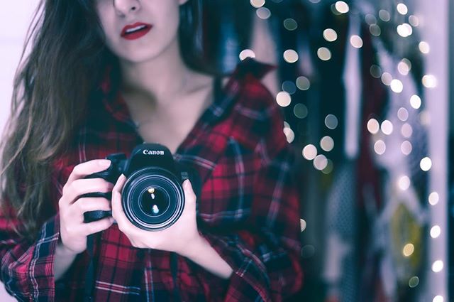 A woman in a flannel shirt holds a Canon camera with a bokeh background, creating a dreamy, intimate portrait.