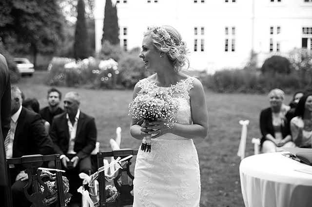 A bride holds her bouquet, smiling during an outdoor wedding ceremony surrounded by guests.