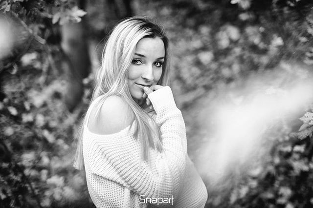 Elegant black and white portrait of a blonde woman in a sweater, set against a softly blurred natural background.