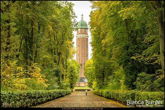 A long pathway leads to a tower in a park, framed by lush green trees and trimmed hedges.