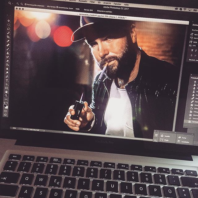A bearded man in a hat vapes while being edited on a MacBook Pro.